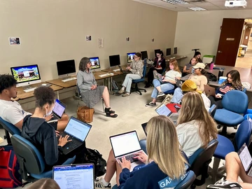 A woman sits with several students recording notes in a class room