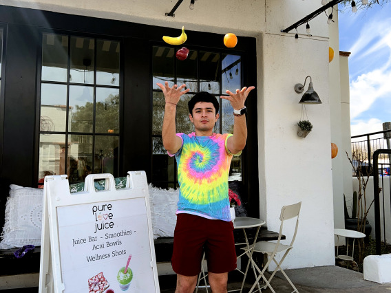 A man juggling fruit in front of a juice bar. 