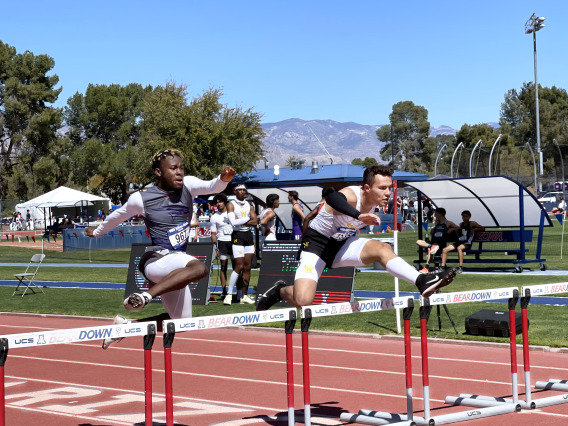 Two high school boys jumping over a hurdle on a track.