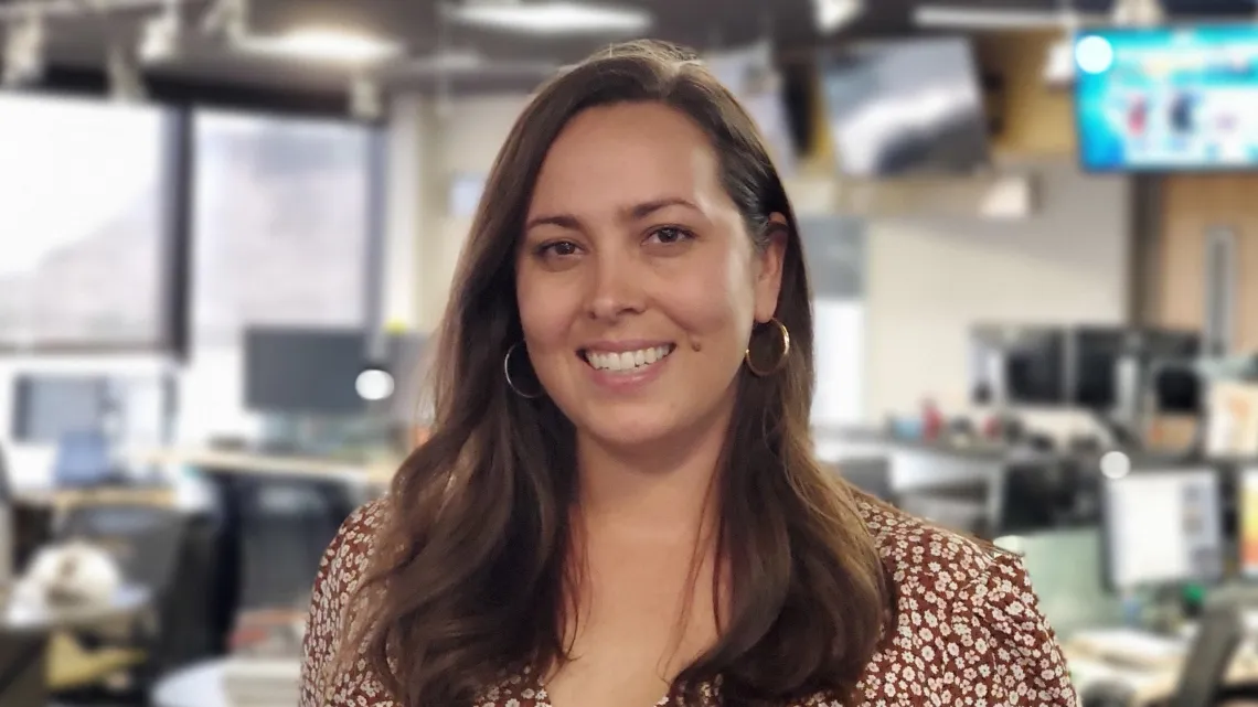 A woman with long brown hair stands in front of an office space.