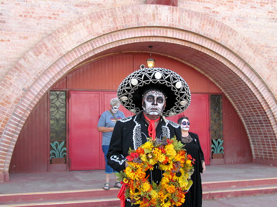 Man in mariachi garb with white face paint holding flower wreath