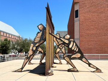 An art sculpture takes up the walkway and depicts four figures pushing or leaning against a border wall-like structure in the middle.