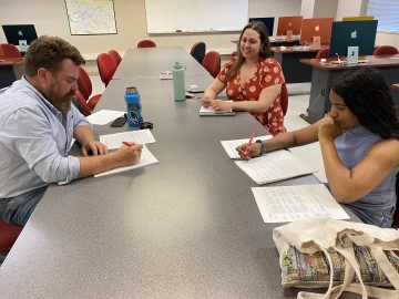 Three people sit at a desk grading papers